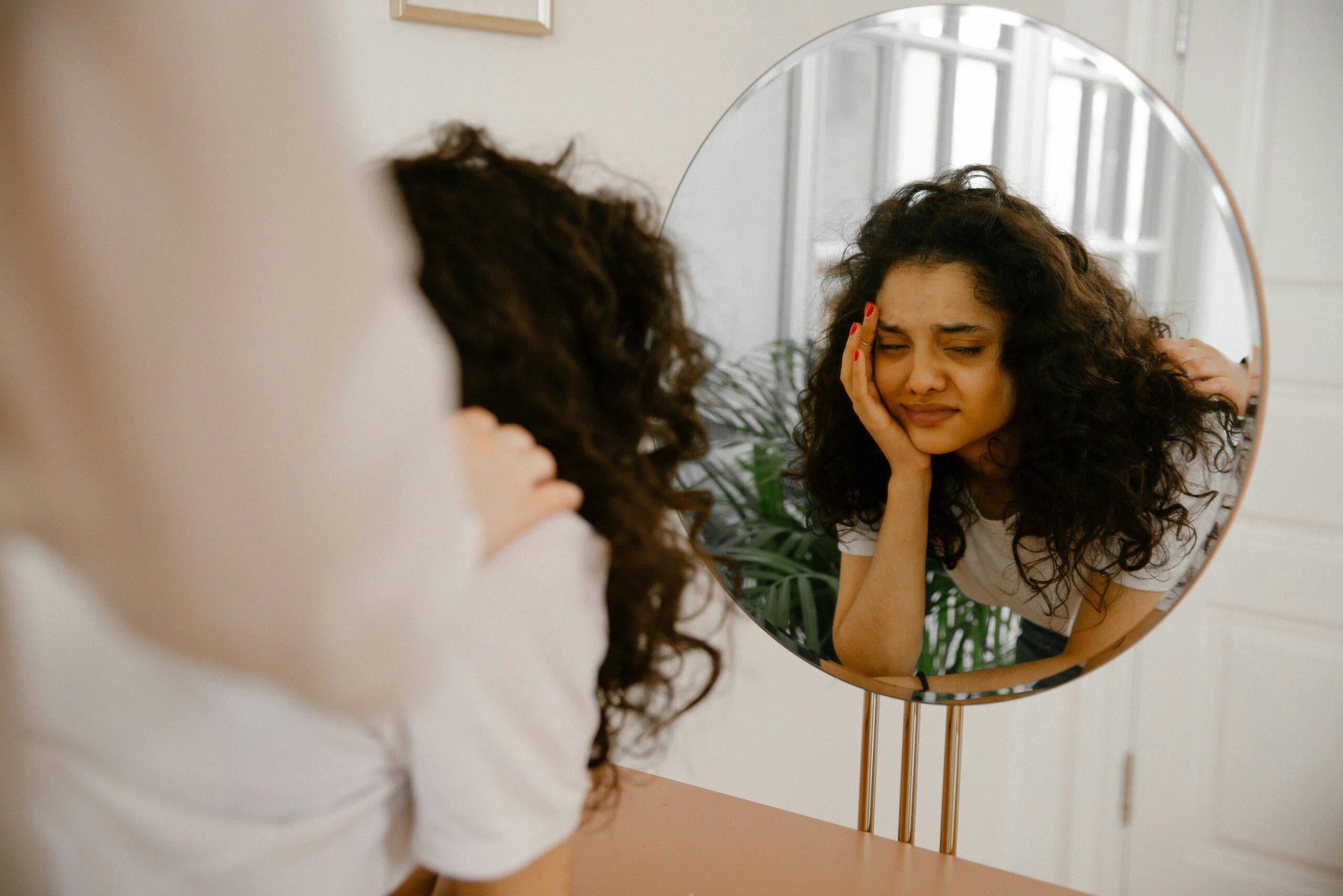 A young woman in front of a mirror expressing self-doubt and contemplation indoors.