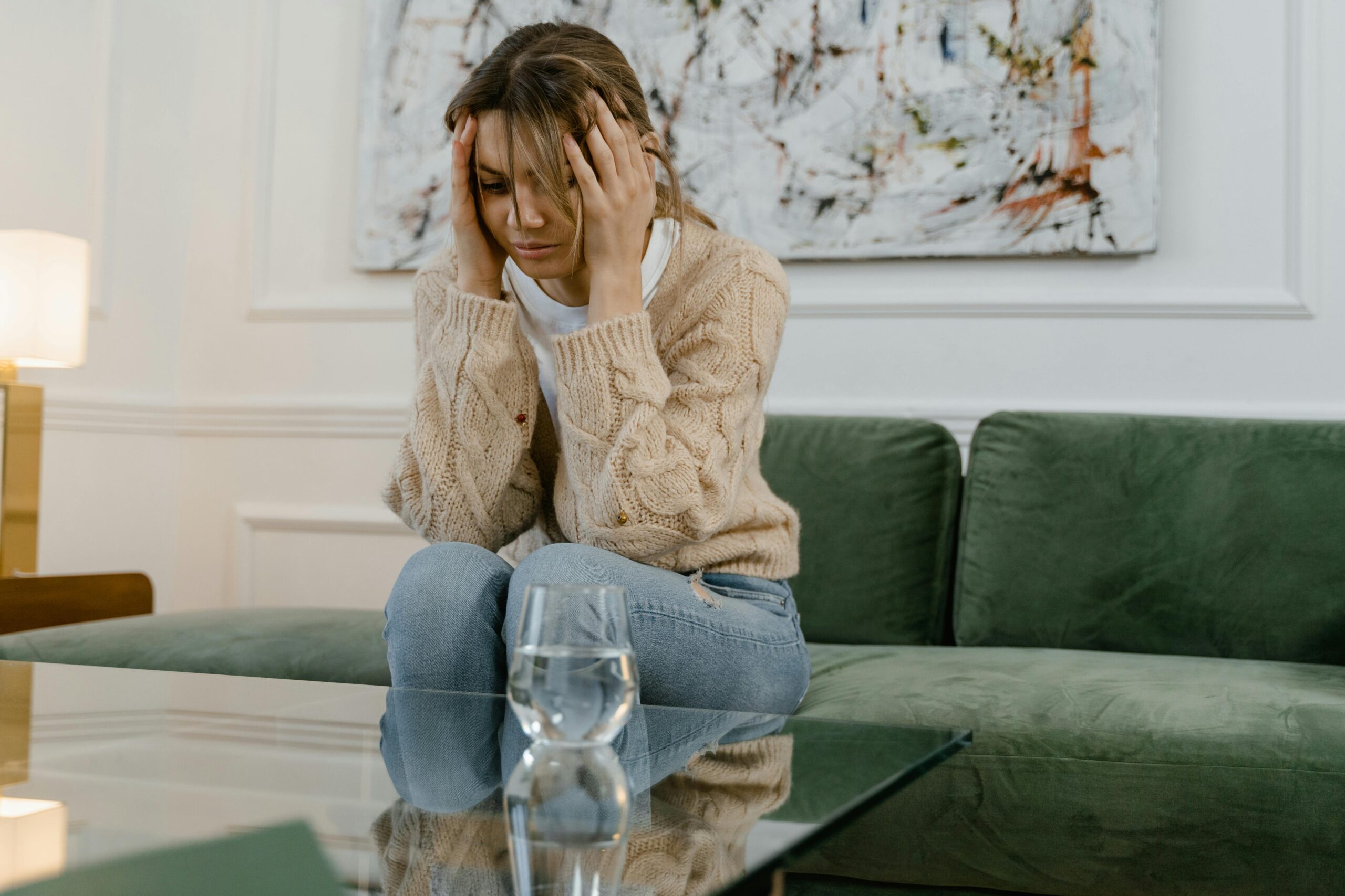 A woman sits on a green sofa looking stressed, holding her head with her hands.